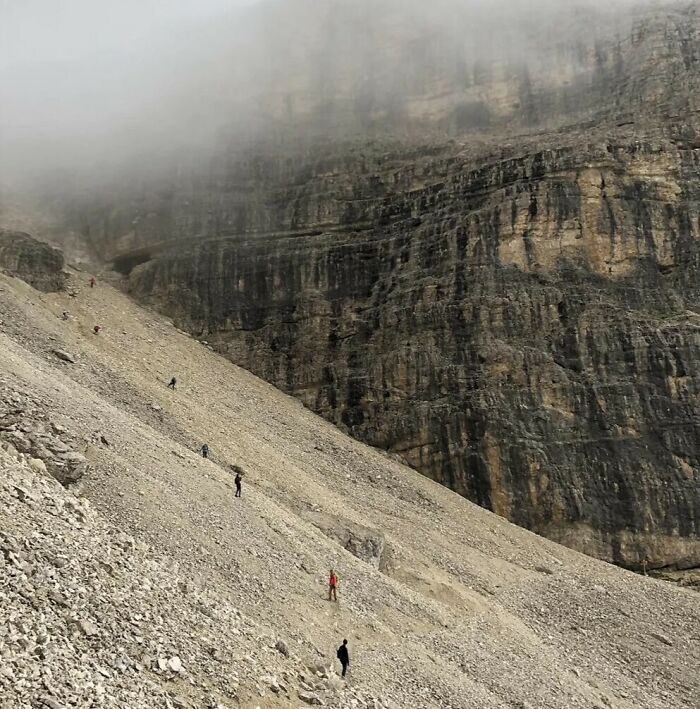 A long line of hikers ascending a steep, rocky mountain slope, emphasizing the true scale of the landscape.