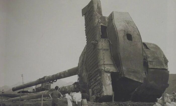 Two people looking at the true scale of a colossal, partially destroyed artillery piece, highlighting its immense size comparison.