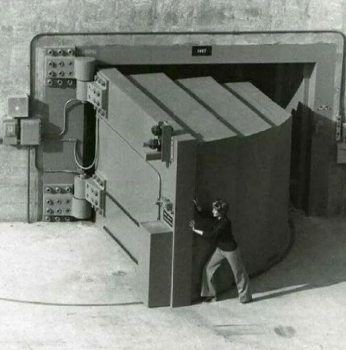 A person standing next to a huge, thick vault door, demonstrating the true scale of its size comparison.