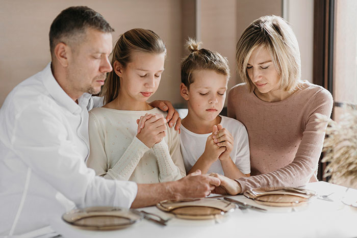 A family of four praying at a dining table, illustrating the complex dynamics of parents raising kids. A family of four praying at a dining table, illustrating the complex dynamics of parents raising kids.