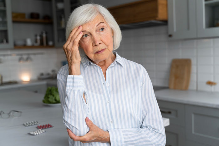 Elderly woman pondering giving up beer for nervous system health, with pills on counter, considering the exact age.