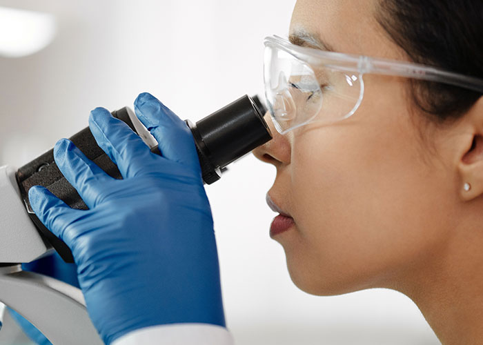 A medical student in safety glasses and blue gloves looks into a microscope, studying human body facts.