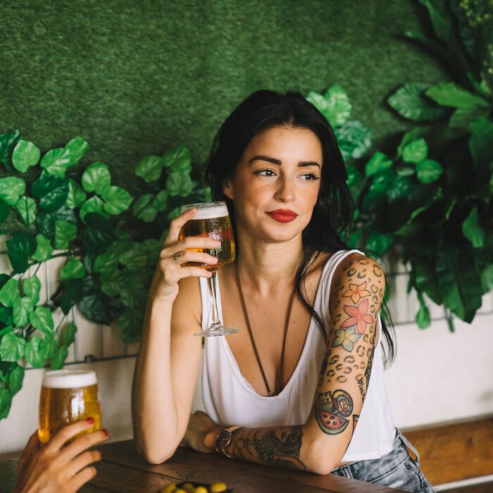 A woman with tattoos holds a glass of beer, sitting at a table with green foliage background. Products, Trends, and Items.