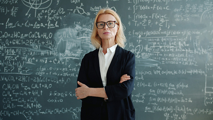 A female teacher with glasses stands in front of a chalkboard filled with equations, learning a lesson. A female teacher with glasses stands in front of a chalkboard filled with equations, learning a lesson.