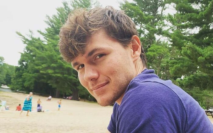 A young man with light brown, messy hair and stubble, wearing a purple shirt, smiles at the camera on a beach with trees in the background, showing his unrecognizable look from Diary of a Wimpy Kid at 28. A young man with light brown, messy hair and stubble, wearing a purple shirt, smiles at the camera on a beach with trees in the background, showing his unrecognizable look from Diary of a Wimpy Kid at 28.