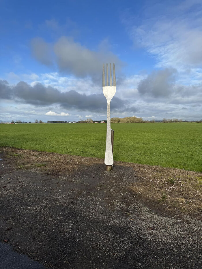 A giant fork sculpture stands in a green field under a cloudy sky, illustrating a visual pun.