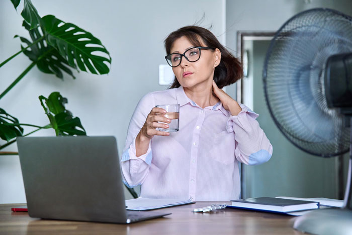 A woman working in the heat, holding a glass of water, with a fan blowing on her. She appears to be uncomfortable in the heat.