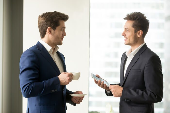 Two male workers chatting in an office, possibly about corporate buzzwords and practices.