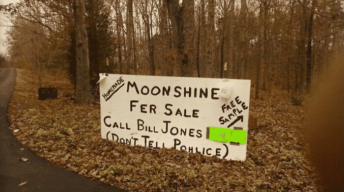 A handwritten sign in a wooded area offering Homemade Moonshine For Sale, a unique photo of the USA.