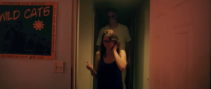 A person with glasses looks over their shoulder at a looming figure in the background of a scary horror movie.
