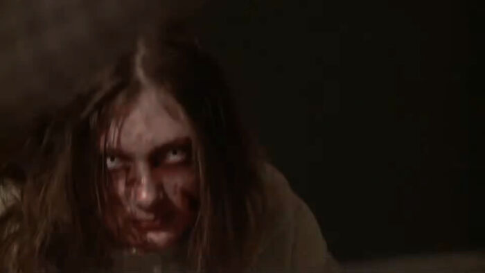 A close-up of a person with long hair, pale eyes, and bloodstains on their face, looking menacingly in a horror movie.