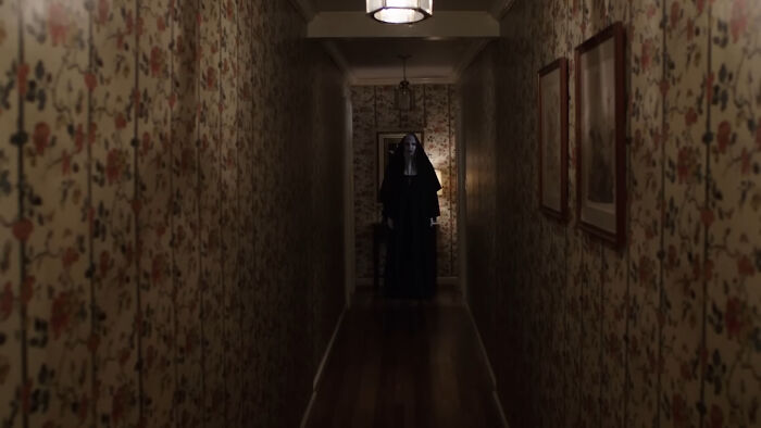 A long, narrow hallway with floral wallpaper leads to a dark, ominous figure in a nun's habit from a horror movie.