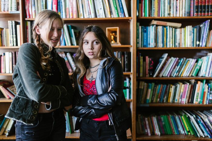 Two girls with long hair stand in a library, looking concerned, realizing they are the problem.