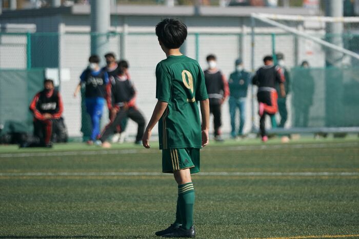 A young boy in a green soccer uniform stands on a field, looking towards other players, realizing he is the problem.