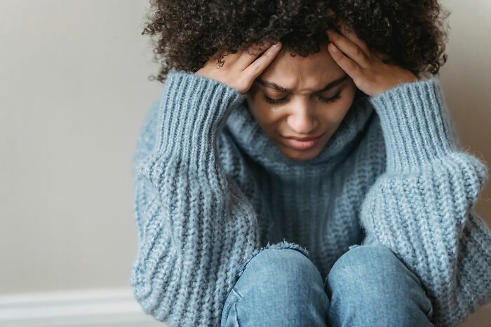 A woman with curly hair, in a blue sweater, sits with her head in her hands, realizing she is the problem.