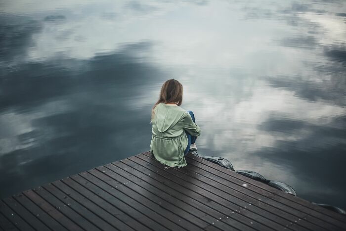 A young woman sits alone on a wooden dock looking out at a calm body of water, realizing she is the problem.