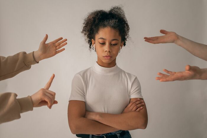A young woman with an unhappy expression, arms crossed, surrounded by gesturing hands, realizing she is the problem.