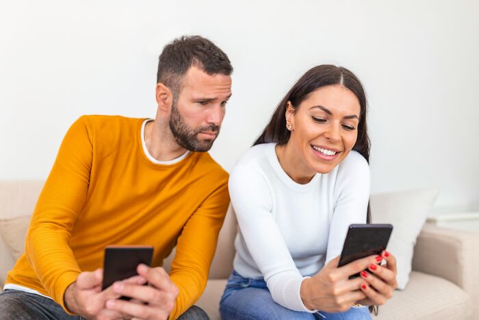 A man looks suspiciously at a woman happily looking at her phone, realizing he is the problem.