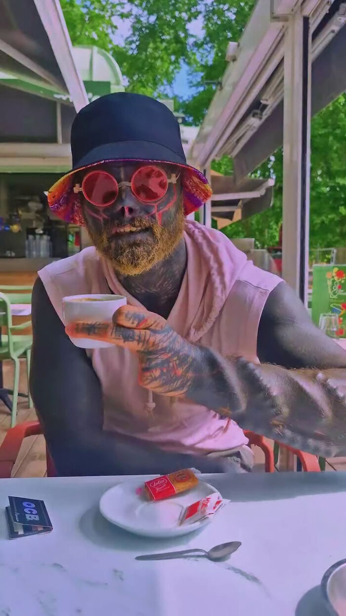 Black Alien man with body modifications, including a removed nose and blackened skin, drinking coffee outdoors, impacting his career. Black Alien man with body modifications, including a removed nose and blackened skin, drinking coffee outdoors, impacting his career.