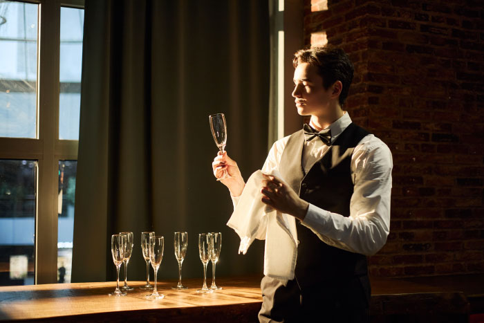 A waiter in a bow tie and vest carefully polishing a champagne glass at a wedding.