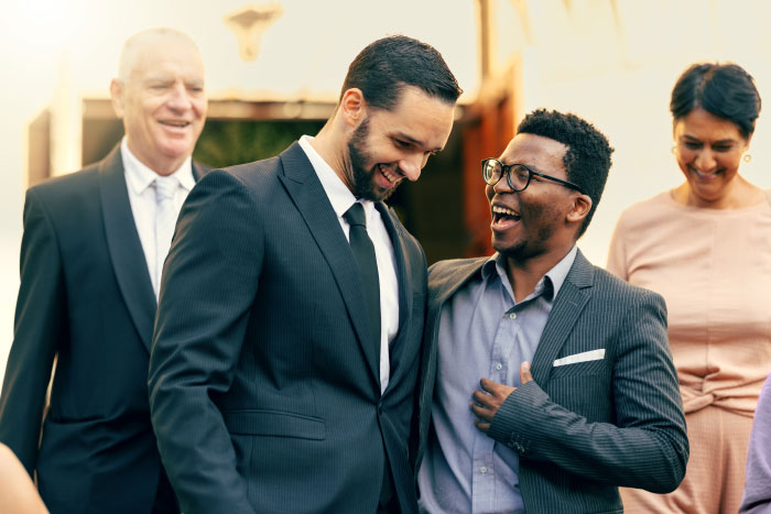 Two men at a wedding laughing heartily, one with an arm around the other, with guests in the background.