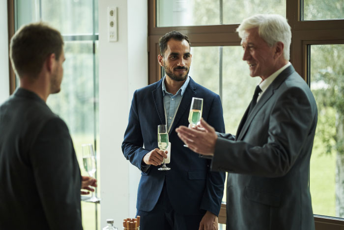 Three men in suits at a wedding, two holding champagne flutes, in conversation.