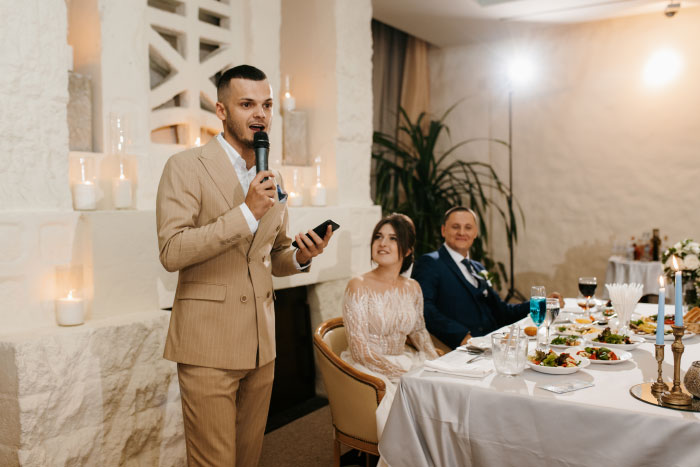 A man in a beige suit gives a speech into a microphone at a wedding reception, a chaotic or memorable wedding moment.