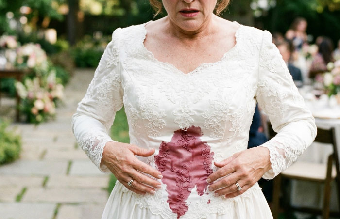 A woman in a white wedding dress looks shocked at a large red wine stain on her gown, a worst thing at a wedding.
