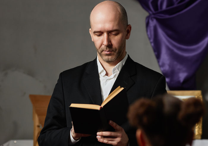 A man in a suit reads from a book, perhaps officiating a wedding where things went chaotic.