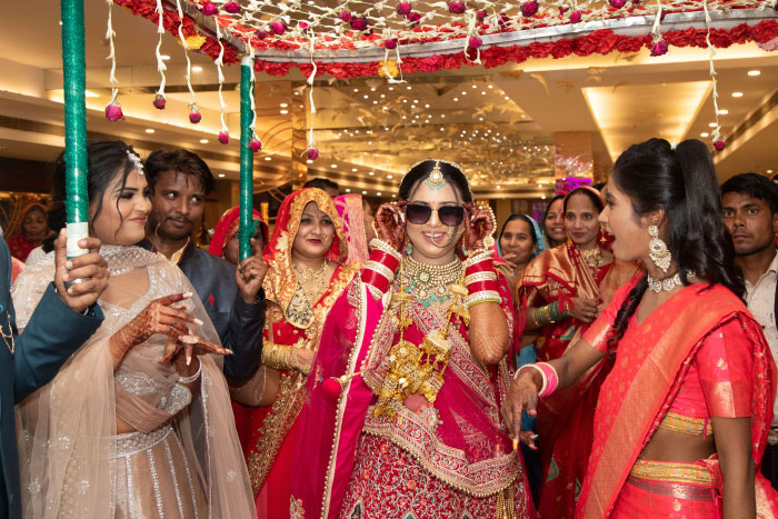 A bride in traditional red attire and sunglasses smiling, being led by people at a chaotic wedding.