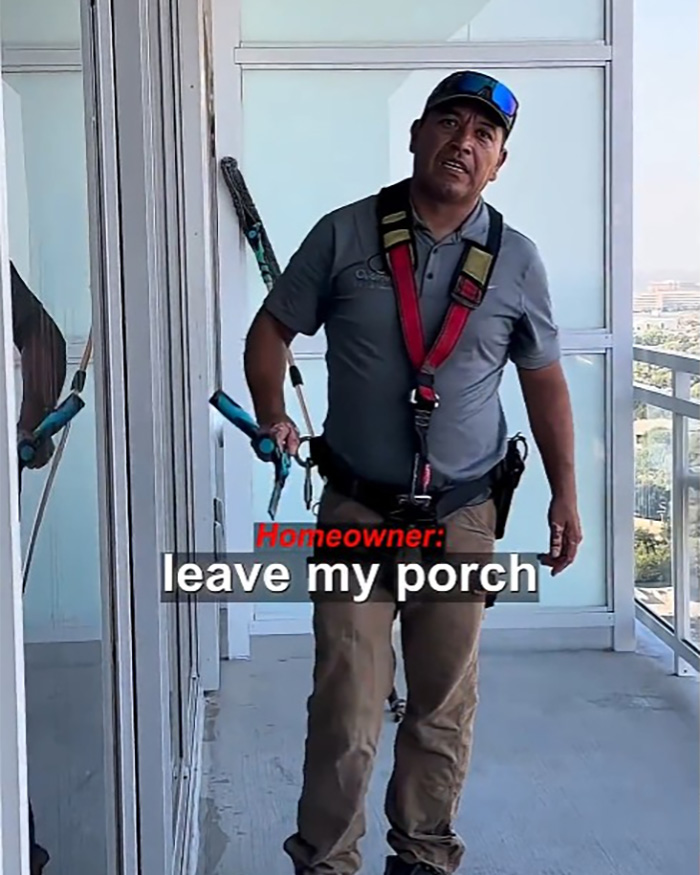 A Hispanic window washer in safety gear stands on a high-rise balcony, looking at the camera with a serious expression. A Hispanic window washer in safety gear stands on a high-rise balcony, looking at the camera with a serious expression.