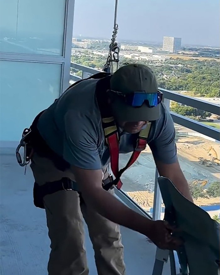 A Hispanic window washer in safety gear bends over on a high-rise balcony, working on his equipment. A Hispanic window washer in safety gear bends over on a high-rise balcony, working on his equipment.