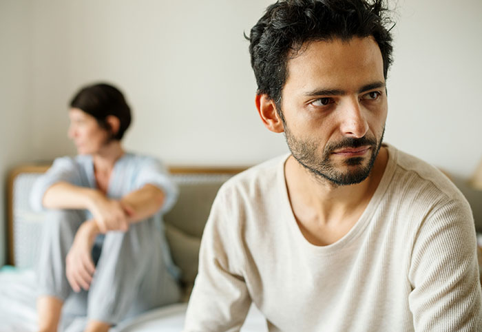 A man with a pensive expression, wife blurred in background. He pressured his wife to have children she never wanted.