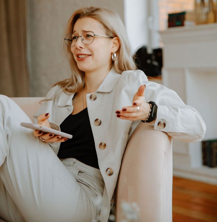 A blonde woman in glasses, looking upset while holding a phone. She's connecting the dots about her BF's trip.