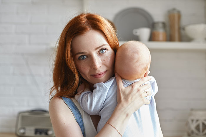 A red-haired woman with freckles holds a baby close to her, looking at the camera. Family and baby.