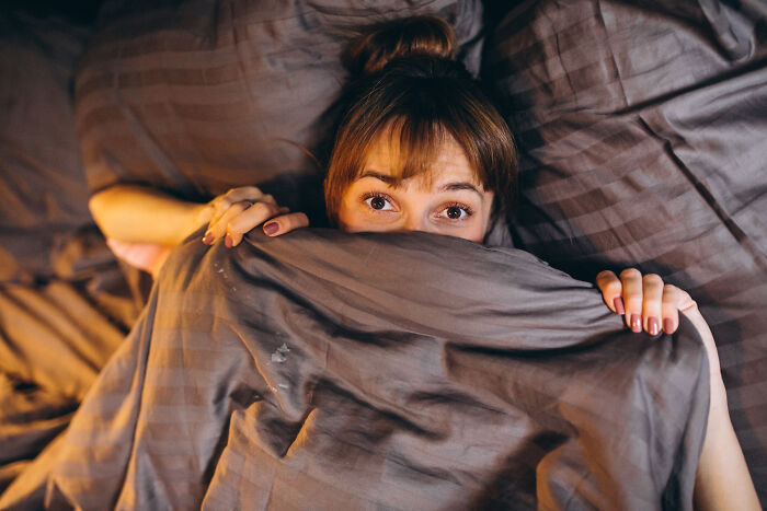 A woman peeking from under a duvet, looking scared, illustrating stories about guests who completely ignored house etiquette.