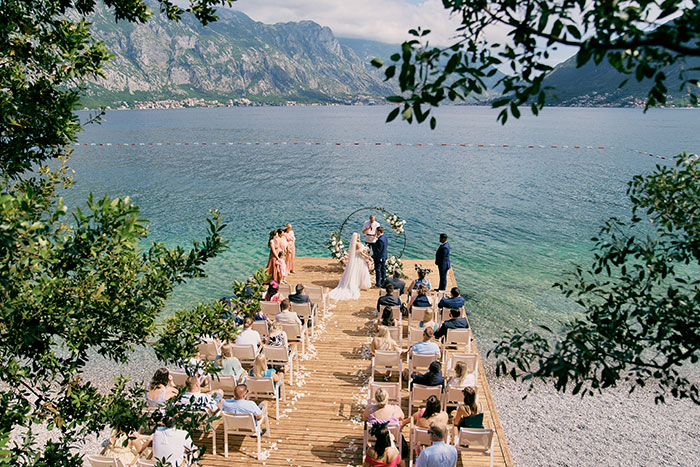 An aerial view of a wedding ceremony on a pier over a lake, surrounded by mountains, highlighting the wedding guest list.