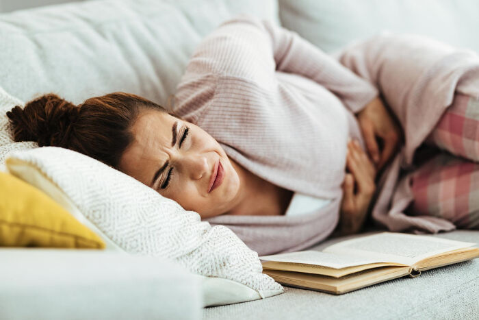 Woman in pain, clutching her stomach on a couch with a book, seeking advice from gynecologists.