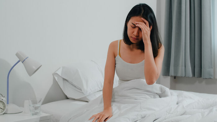 A woman sits on her bed looking distressed, holding her head, reflecting the important advice from gynecologists.
