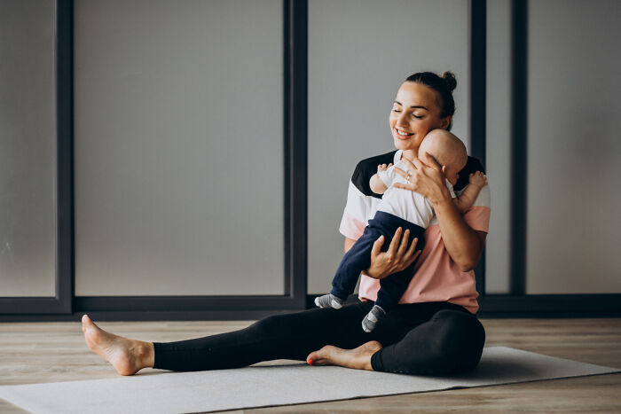 A mother sits on a yoga mat, smiling and holding her baby, emphasizing women's health and family.