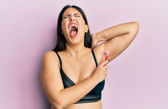 A woman in a black bra winces while shaving her underarm, highlighting common women's health concerns.