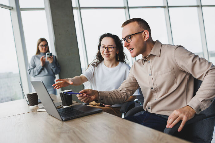 Two workers collaborate at a laptop, discussing corporate buzzwords and practices in a bright office.