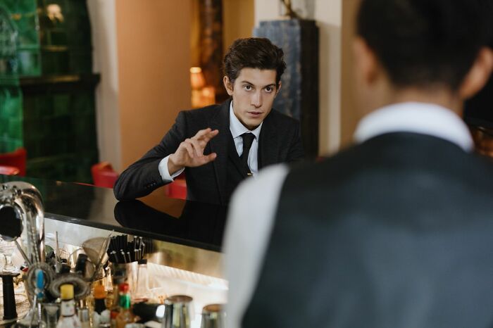 A man in a suit talking to a bartender, possibly displaying first date red flags with his behavior.