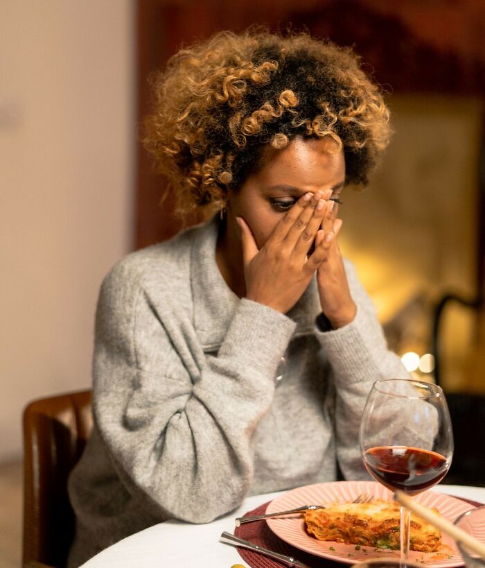 A woman covers her face at a table with food and wine, showing distress from first date red flags.