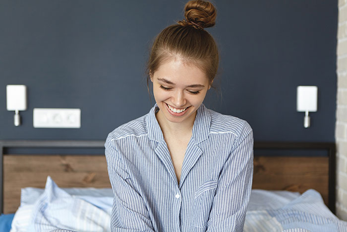 A smiling teen woman in striped pajamas, with hair in a bun, sits on a bed, embodying freedom from a controlling mom. A smiling teen woman in striped pajamas, with hair in a bun, sits on a bed, embodying freedom from a controlling mom.