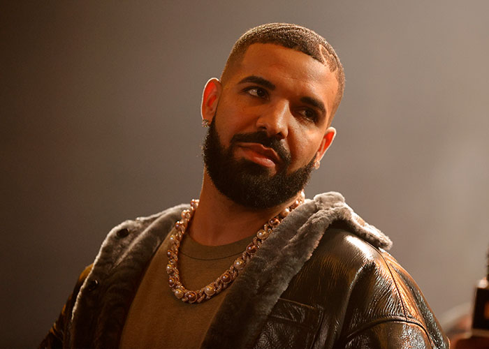 Drake looking serious, wearing a pearl chain and fur-lined jacket, related to his dating challenge.
