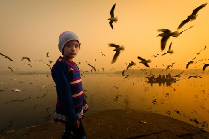 A young child standing by a river, looking directly at the viewer, with many birds flying overhead. Murat Bakmaz captures the beauty of everyday life.