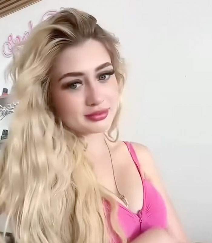 A beauty influencer with long blonde hair, wearing a pink top, looking at the camera during a livestream. A beauty influencer with long blonde hair, wearing a pink top, looking at the camera during a livestream.