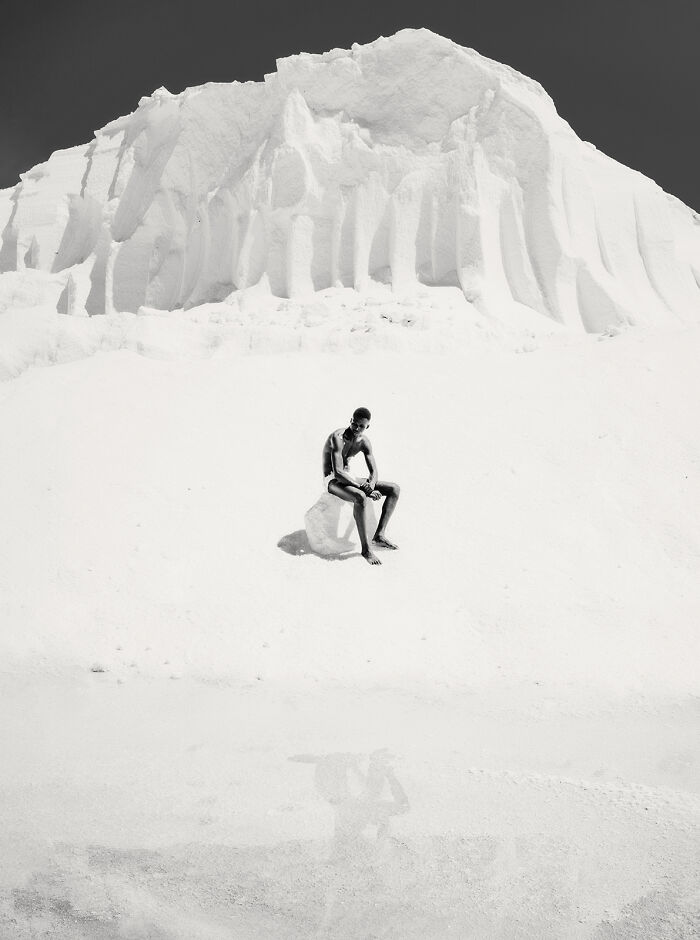 Stark minimalist photography of a person sitting on a small object in a vast, white, snow-like landscape.