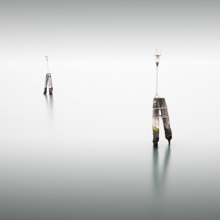 Minimalist photography of a wooden pier structure emerging from calm, misty water under a soft, white sky.